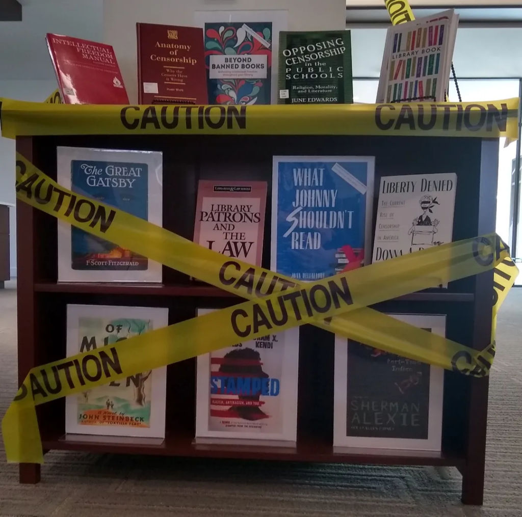 Yellow CAUTION tape across a bookshelf containing titles such as "Anatomy of Censorship" "Opposing Censorship in the Public Schools" "The Great Gatsby" "Library Patrons and the Law" and "Liberty Denied"
